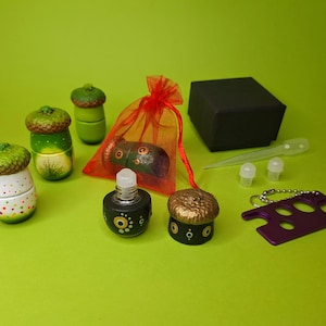 May include: A set of six acorn-shaped bottles with various designs, including green, gold, and black with white polka dots. The bottles are arranged on a green surface with a red mesh bag, a black box, and a purple tool.