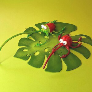 May include: Two whimsical, cartoon-like bugs with large eyes and long snouts are perched on a green leaf. One bug is green and the other is red.