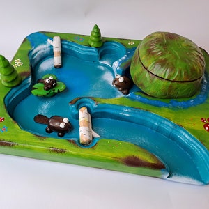 May include: A wooden game board with a blue river, green grass, and brown beavers. The board is decorated with flowers and mushrooms. There are two logs in the river.