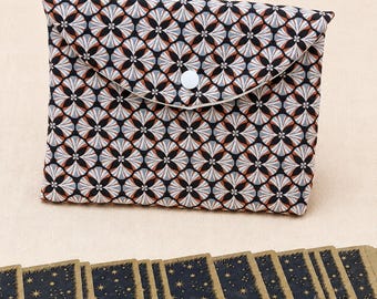 May include: A fabric pouch featuring a repeating geometric pattern in black, white, and rust orange with a white snap closure, positioned above a row of rectangular cards with a dark blue celestial star print and gold-coloured edges.