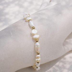 Könnte beinhalten: Ein zartes Armband mit unregelmäßig geformten weißen Perlen und kleinen goldfarbenen Perlen. Die Perlen sind aufgereiht und bilden ein raffiniertes und elegantes Schmuckstück. Das Armband wird auf einer weißen Oberfläche präsentiert.