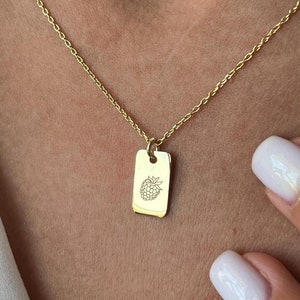 May include: A gold necklace with a rectangular pendant showing a raspberry design. The pendant is suspended from a fine chain. The necklace is worn around the neck. The image is a close-up.
