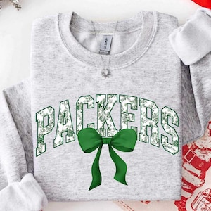 Könnte beinhalten: Ein hellgraues Sweatshirt mit Rundhalsausschnitt, das das Wort PACKERS in Blockschrift mit einem grün-weißen Blumenmuster zeigt. Direkt unter dem Text befindet sich eine einfarbige grüne Schleife. Das Kleidungsstück hat lange Ärmel.