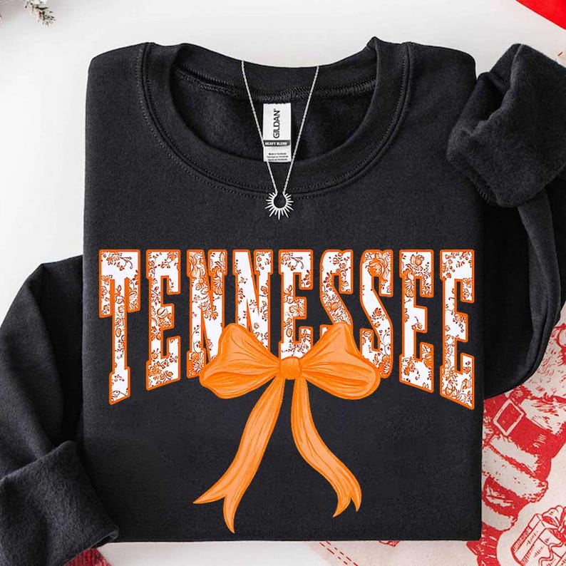 K&ouml;nnte beinhalten: Ein schwarzes Langarm-Sweatshirt mit dem Wort TENNESSEE in wei&szlig;en Blockbuchstaben, die mit einem orangefarbenen Blumenmuster gef&uuml;llt sind. Eine gro&szlig;e, einfarbige orangefarbene Schleife befindet sich direkt unter der Wortmitte.