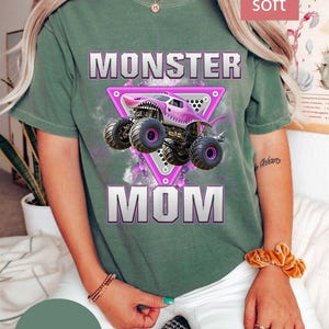 May include: Moss green t-shirt with a pink monster truck graphic inside a pink triangle, the words "MONSTER MOM" are printed below the truck. The shirt also has the words "super soft" printed on it.