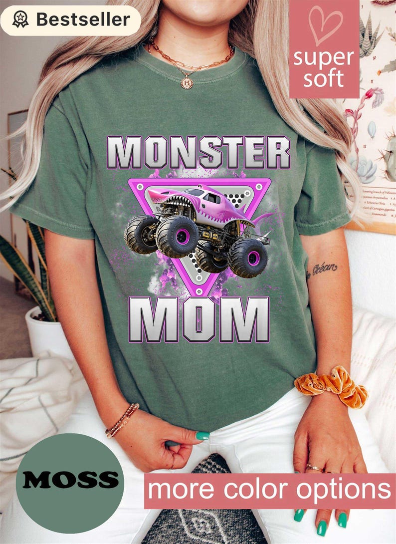 May include: Moss green t-shirt with a pink monster truck graphic inside a pink triangle, the words "MONSTER MOM" are printed below the truck. The shirt also has the words "super soft" printed on it.