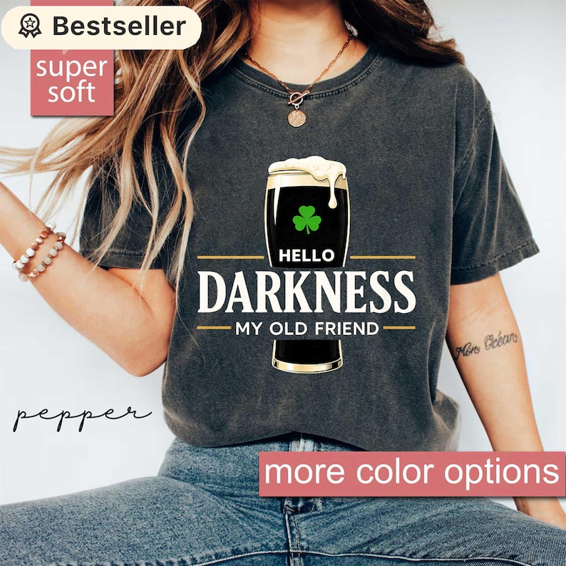 May include: Dark gray t-shirt featuring a graphic of a pint of stout with a shamrock and the text "HELLO DARKNESS MY OLD FRIEND." The shirt has a vintage look and is labeled "Bestseller" and "super soft."