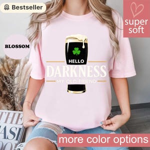 May include: A light pink t-shirt featuring a graphic of a dark stout with a shamrock and the text "HELLO DARKNESS MY OLD FRIEND." The shirt has a "super soft" label and a "Bestseller" tag.