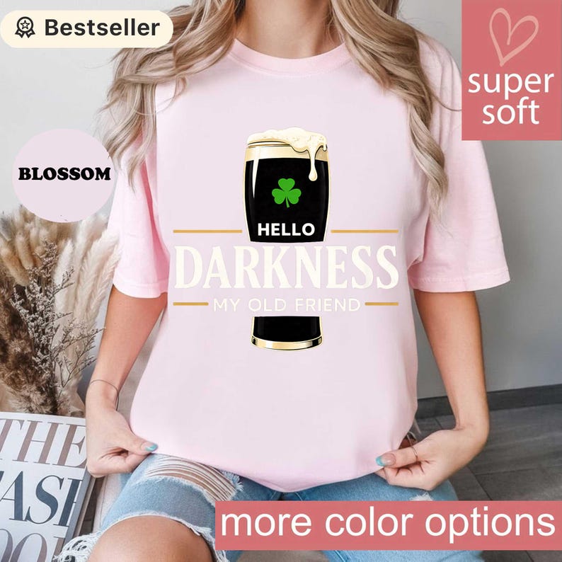 May include: A light pink t-shirt featuring a graphic of a dark stout with a shamrock and the text "HELLO DARKNESS MY OLD FRIEND." The shirt has a "super soft" label and a "Bestseller" tag.