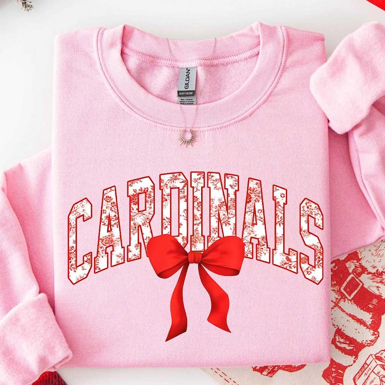 May include: A light pink crewneck sweatshirt featuring the word CARDINALS in a white block font with a red floral pattern. A large, bright red bow is positioned directly below the text, centered on the chest of the garment.