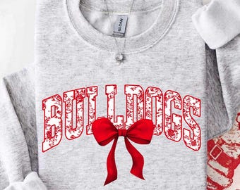 May include: A light gray crewneck sweatshirt featuring the word BULLDOGS in large, red, floral-patterned block letters across the chest. A prominent, solid red bow is positioned centrally over the text, and a silver necklace is visible at the neck.