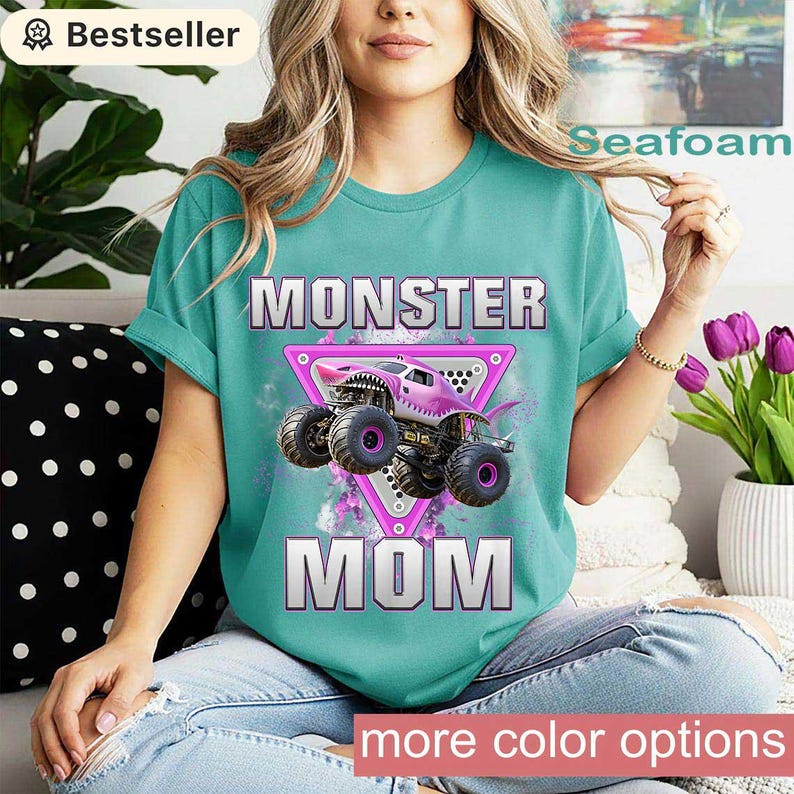 May include: Seafoam green t-shirt with the words "MONSTER MOM" and a pink monster truck graphic. The graphic is inside a pink triangle. The shirt has rolled-up sleeves and is worn by a person.