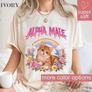 May include: Ivory-colored t-shirt with the text "ALPHA MALE" in pink, featuring a cartoon orange tabby cat with blue eyes, a rainbow, flowers, and butterflies. The shirt also has the text "more color options".