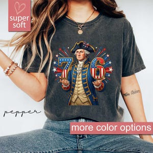 Op de afbeelding: Donkergrijs t-shirt met een afbeelding van George Washington met de cijfers 76, met rode, witte en blauwe accenten. Het shirt heeft de woorden "super soft" en "pepper" erop gedrukt, met een banner "more color options".