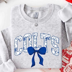 Könnte beinhalten: Ein hellgraues Sweatshirt mit Rundhalsausschnitt, das das Wort COLTS in großen Blockbuchstaben mit einem blau-weißen Blumenmuster zeigt. Eine einfarbige blaue Schleife ist direkt unter dem Text platziert. Eine silberne Kette mit Anhänger ist sichtbar.