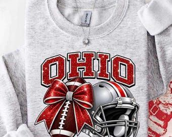 May include: A light gray crewneck sweatshirt featuring the word OHIO in large red glittery block letters. Below the text is a brown football adorned with a large red glittery bow, positioned next to a silver and red football helmet.