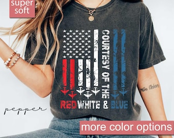 May include: Dark gray t-shirt featuring a stylized American flag design. The flag incorporates red, white, and blue stripes with the text "COURTESY OF THE RED WHITE & BLUE" and airplane silhouettes. The shirt has the text "super soft" and "more color options".