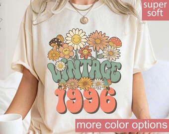May include: Ivory-colored t-shirt with a retro floral design and the text "VINTAGE 1996." The graphic features smiling flowers, mushrooms, and the words in a groovy font. The shirt has short sleeves and a relaxed fit.