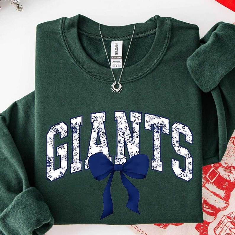 May include: A dark forest green crewneck sweatshirt featuring the word GIANTS in large, white block letters with a blue floral pattern. A solid navy blue bow graphic is positioned below the text. A silver sunburst pendant necklace is visible.