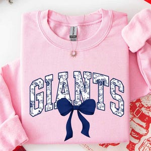 May include: A light pink crewneck sweatshirt featuring the word GIANTS in a white and blue floral pattern across the chest. A solid dark blue bow is positioned directly below the text. The garment has a ribbed collar and long sleeves.