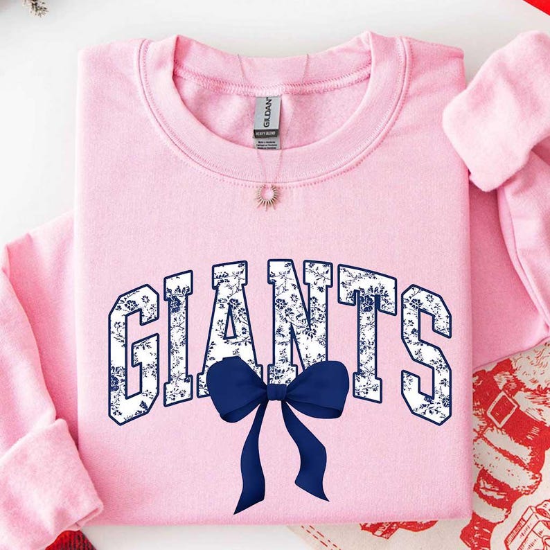 May include: A light pink crewneck sweatshirt featuring the word GIANTS in a white and blue floral pattern across the chest. A solid dark blue bow is positioned directly below the text. The garment has a ribbed collar and long sleeves.