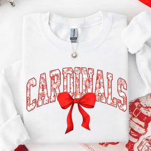 May include: A white long-sleeve sweatshirt featuring the word CARDINALS in a red and white floral pattern across the chest. A large, solid red bow is positioned directly below the text. A silver necklace with a small pendant rests at the neckline.