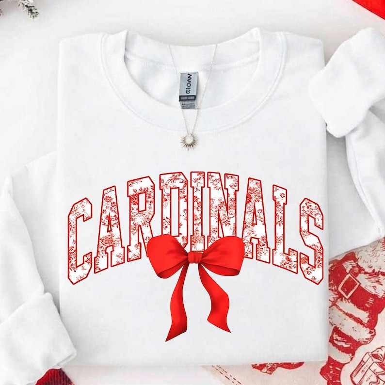 May include: A white long-sleeve sweatshirt featuring the word CARDINALS in a red and white floral pattern across the chest. A large, solid red bow is positioned directly below the text. A silver necklace with a small pendant rests at the neckline.