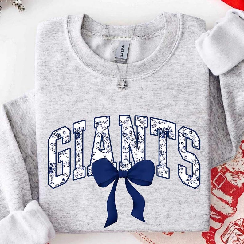 May include: A light gray heather crewneck sweatshirt featuring the word GIANTS in large block letters with a blue and white floral pattern. A solid dark blue bow is positioned directly below the center of the text on the front of the garment.