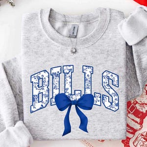 Könnte beinhalten: Ein hellgraues Sweatshirt mit Rundhalsausschnitt, das das Wort BILLS in großen Blockbuchstaben mit einem blau-weißen Blumenmuster zeigt. Eine einfarbige blaue Schleife ist an der Basis der Buchstaben angebracht. Eine silberne Kette mit Anhänger ist sichtbar.