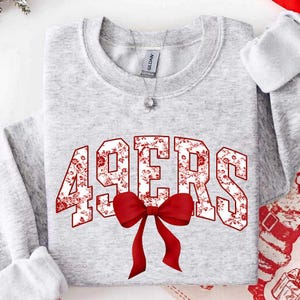 Könnte beinhalten: Ein hellgraues Sweatshirt mit Rundhalsausschnitt und dem Schriftzug 49ERS in einer fetten Blockschrift, die mit einem rot-weißen Blumenmuster gefüllt ist. Eine große, einfarbige rote Schleife befindet sich direkt unter der Mitte der Grafik.