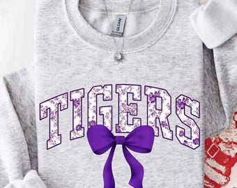 May include: A light gray crewneck sweatshirt featuring the word TIGERS in large block letters with a purple floral pattern. A solid purple bow is positioned directly below the text. The garment has a ribbed collar and long sleeves.