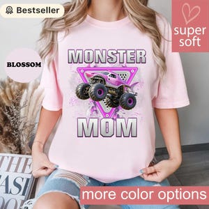 May include: Light pink t-shirt with the words "MONSTER MOM" in silver block letters. A pink monster truck graphic is centered within a pink triangle. The shirt has the words "super soft" and "more color options" on it.