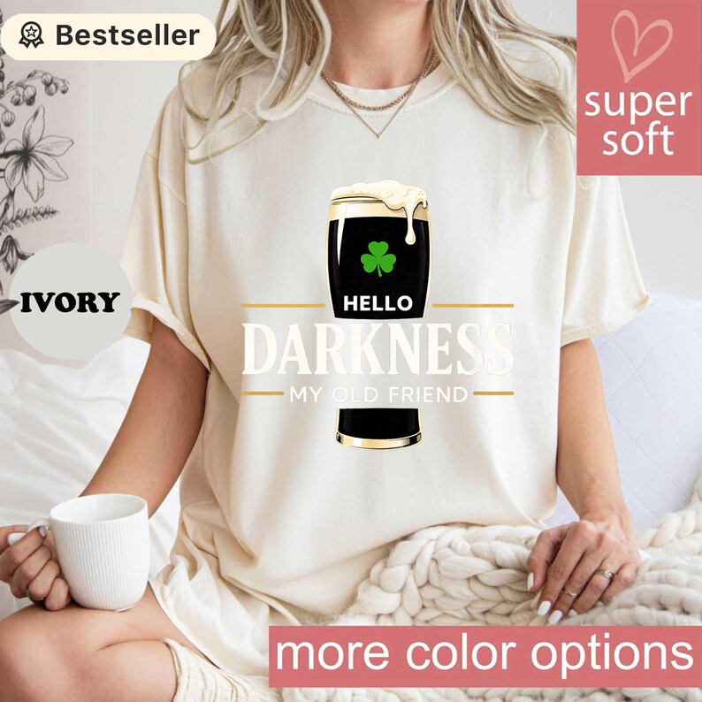 May include: Ivory-colored t-shirt featuring a graphic of a stout beer with a shamrock and the words "Hello Darkness My Old Friend." The shirt has a "super soft" label and the words "more color options" are visible.