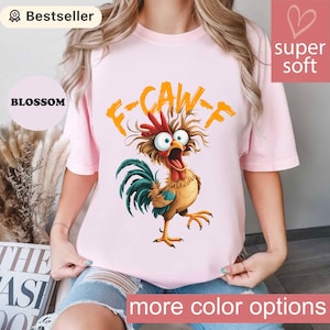 May include: A pale pink t-shirt with a cartoon rooster design and the orange text "F-CAW-F". The rooster has a red comb, brown feathers, and a blue and green tail. The shirt also has the text "more color options".