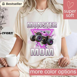 May include: Ivory-colored t-shirt featuring a pink monster truck graphic with the words "MONSTER MOM" in a bold, outlined font. The design includes a triangle frame and a "super soft" label.
