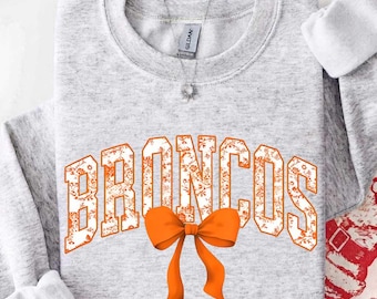 May include: A light gray crewneck sweatshirt featuring the word BRONCOS in large, white block letters with an orange floral pattern. A solid orange bow is positioned directly below the text. A silver necklace with a small pendant is visible.