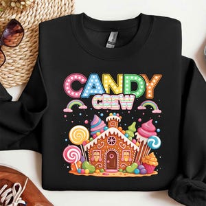 Könnte beinhalten: Schwarzes Sweatshirt mit einem farbenfrohen "CANDY CREW" -Aufdruck. Das Design zeigt ein Lebkuchenhaus, umgeben von Süßigkeiten, Lollis und Cupcakes. Regenbogen und farbenfrohe Akzente runden das süße Thema ab.