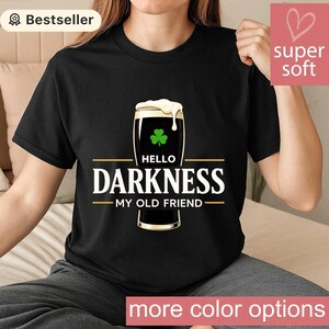 May include: Black t-shirt featuring a pint of stout with a shamrock and the text "Hello Darkness My Old Friend." The shirt has a "Bestseller" tag and a "super soft" label. The words "more color options" are at the bottom.
