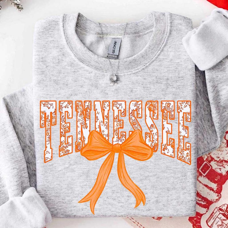 K&ouml;nnte beinhalten: Ein hellgraues Sweatshirt mit Rundhalsausschnitt, auf dessen Brust das Wort TENNESSEE in einem wei&szlig;-orangen Blumenmuster gedruckt ist. Eine gro&szlig;e, einfarbig orangefarbene Schleifengrafik liegt mittig &uuml;ber dem unteren Teil des Textes.