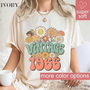 May include: Ivory-colored t-shirt with a retro floral design and the text "VINTAGE 1966." The graphic features smiling daisies, mushrooms, and other flowers in shades of orange, green, and yellow. The shirt has short sleeves and a relaxed fit.