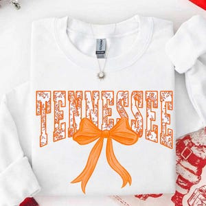 K&ouml;nnte beinhalten: Ein wei&szlig;es Langarm-Sweatshirt mit dem Wort TENNESSEE in gro&szlig;en Blockbuchstaben mit orangefarbenem Blumenmuster. Eine einfarbige orangefarbene Schleife ist mittig unter dem Text platziert. Eine silberne Halskette mit kleinem Anh&auml;nger ist zu sehen.