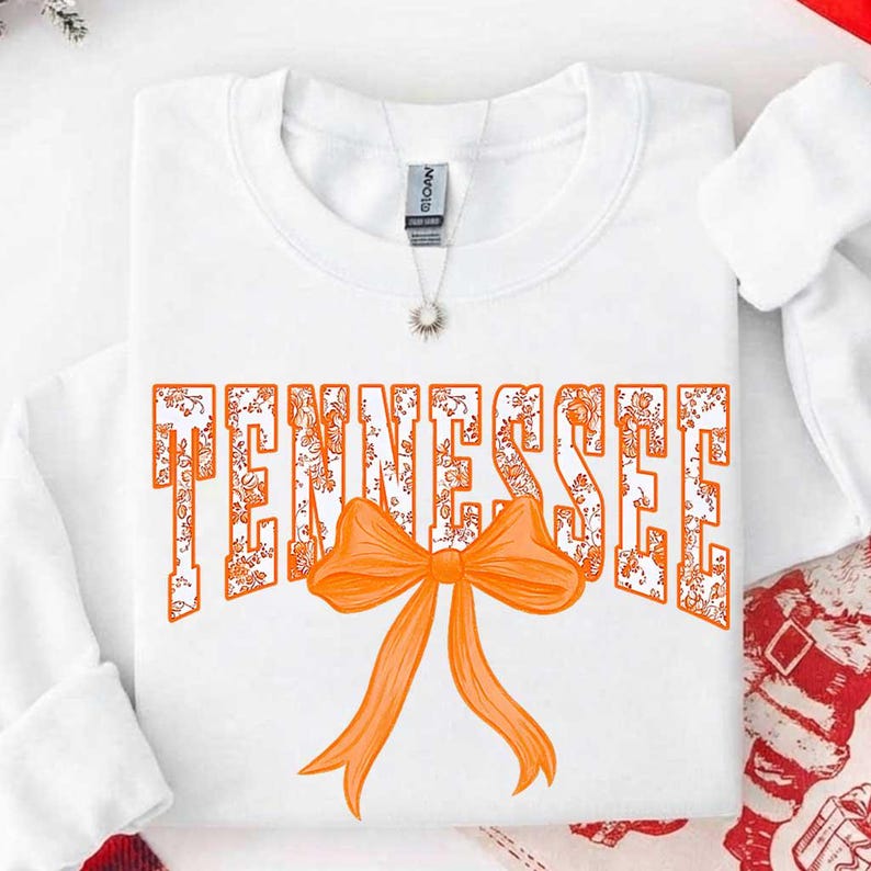 K&ouml;nnte beinhalten: Ein wei&szlig;es Langarm-Sweatshirt mit dem Wort TENNESSEE in gro&szlig;en Blockbuchstaben mit orangefarbenem Blumenmuster. Eine einfarbige orangefarbene Schleife ist mittig unter dem Text platziert. Eine silberne Halskette mit kleinem Anh&auml;nger ist zu sehen.