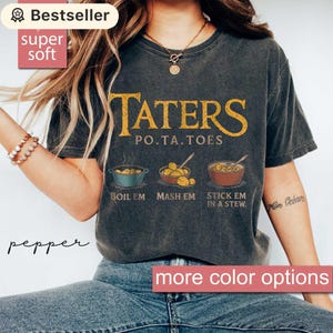 May include: Dark gray t-shirt with the text "TATERS PO.TA.TOES" in gold lettering. Below the text are illustrations of potatoes being boiled, mashed, and in a stew. The shirt also has the text "BOIL EM MASH EM STICK EM IN A STEW."