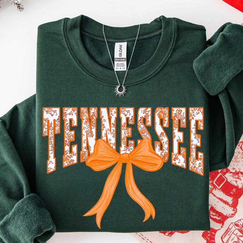 K&ouml;nnte beinhalten: Ein dunkelgr&uuml;nes Sweatshirt mit Rundhalsausschnitt und dem Wort TENNESSEE in wei&szlig;en Blockbuchstaben, die mit einem orangefarbenen Blumenmuster gef&uuml;llt sind. Eine gro&szlig;e, einfarbige orangefarbene Schleife befindet sich unter dem Schriftzug.