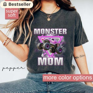 May include: Dark grey t-shirt with the words "MONSTER MOM" in white and pink. A pink monster truck graphic is centered on the shirt. The shirt has a "Bestseller" and "super soft" label.