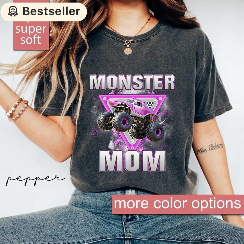 May include: Dark grey t-shirt with the words "MONSTER MOM" in white and pink. A pink monster truck graphic is centered on the shirt. The shirt has a "Bestseller" and "super soft" label.