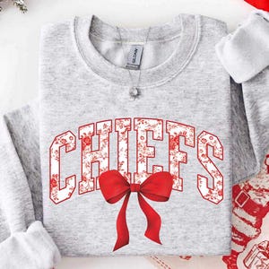 Könnte beinhalten: Ein hellgraues Sweatshirt mit Rundhalsausschnitt, das das Wort CHIEFS in großen Blockbuchstaben mit einem rot-weißen Blumenmuster zeigt. Eine auffällige, einfarbig rote Schleife befindet sich direkt unter der Mitte der Grafik.