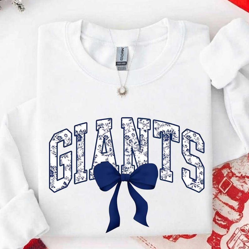 May include: A white long-sleeve sweatshirt featuring the word GIANTS in a blue and white floral pattern. A solid dark blue bow is positioned directly below the text. The garment has a crew neckline and a visible brand tag at the inner collar.