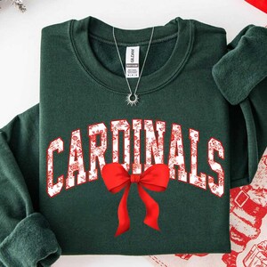 May include: A dark forest green crewneck sweatshirt featuring the word CARDINALS in white block letters with a red floral pattern. A bright red bow graphic is positioned below the text. A silver sunburst pendant necklace rests on the neckline.