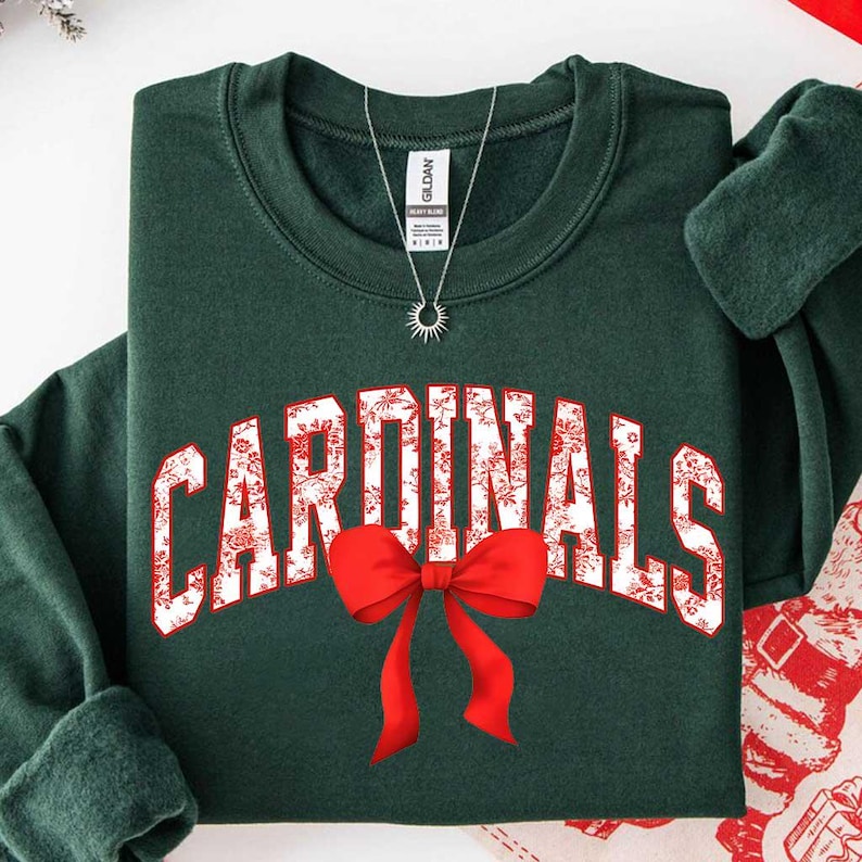 May include: A dark forest green crewneck sweatshirt featuring the word CARDINALS in white block letters with a red floral pattern. A bright red bow graphic is positioned below the text. A silver sunburst pendant necklace rests on the neckline.